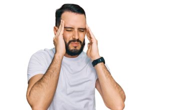 Sakallı, sıradan beyaz tişörtlü, eli başının üstünde genç bir adam. Stres yüzünden baş ağrısı çekiyor. migren ağrısı. 