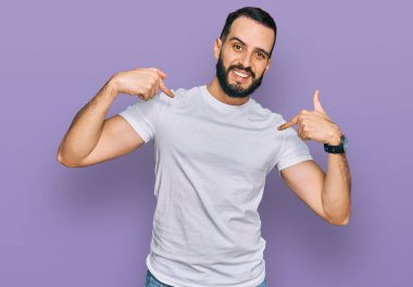 Sakallı, sıradan beyaz tişörtlü, kendine güvenen, gülümseyen, kendini gururlu ve mutlu gösteren genç bir adam.. 