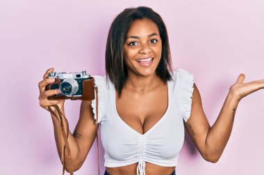 Genç Afrikalı Amerikalı kız elinde klasik kamerayla başarıyı mutlu bir gülümsemeyle ve kazanan yüz ifadesiyle kutluyor. 