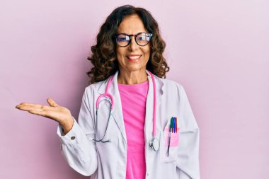 Orta yaşlı İspanyol kadın doktor üniforması giyiyor ve gözlüklü neşeli bir şekilde gülümsüyor ve avuç içi kameraya bakıyor.. 