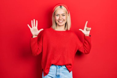 Güzel sarışın kadın gündelik kırmızı süveter giyer ve yedinci parmaklarıyla işaret ederken kendine güvenen ve mutlu bir şekilde gülümser.. 