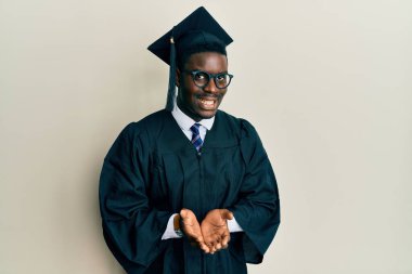 Mezuniyet şapkası ve tören cübbesi takan yakışıklı siyahi bir adam elleri avuçlarında gülümseyen ya da el hareketi yapan. Bekleme ve koruma 