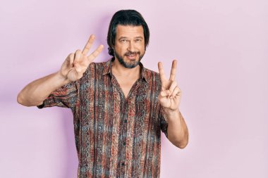 Orta yaşlı, günlük kıyafetler giyen, gülümseyen, parmaklarını zafer işareti yaparken gösteren beyaz bir adam. İki numara.. 