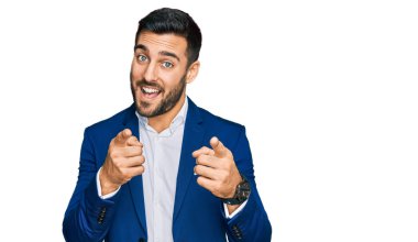 İş ceketi giyen genç İspanyol adam mutlu ve komik yüzlü parmaklarını kameraya doğrultuyor. iyi enerji ve titreşimler. 