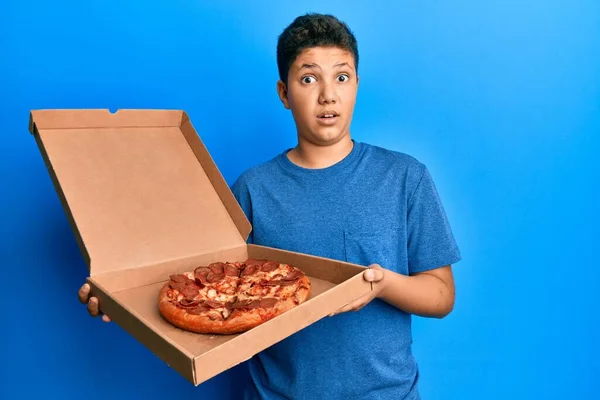 十代のヒスパニック少年食べるおいしいペパロニピザで衝撃的な顔 懐疑的で皮肉を探して オープン口で驚いて — ストック写真