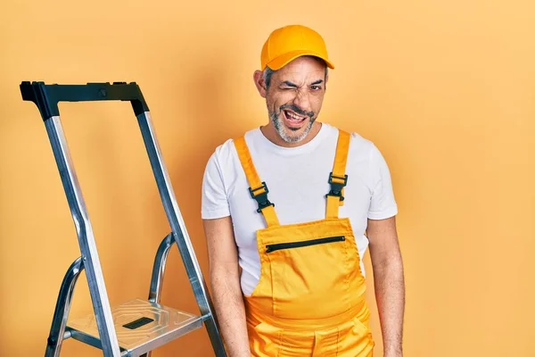Handsome middle age man with grey hair holding ladder winking looking at the camera with sexy expression, cheerful and happy face.
