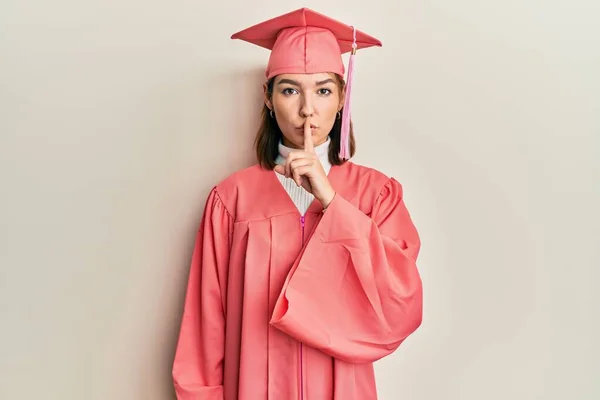 卒業キャップと儀式用のローブを着た若い白人女性が唇に指で静かにするように求めています 沈黙と秘密の概念 — ストック写真