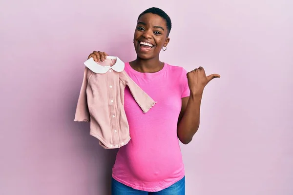 Jovem Afro Americana Esperando Bebê Segurando Roupas Apontando Polegar Para — Fotografia de Stock