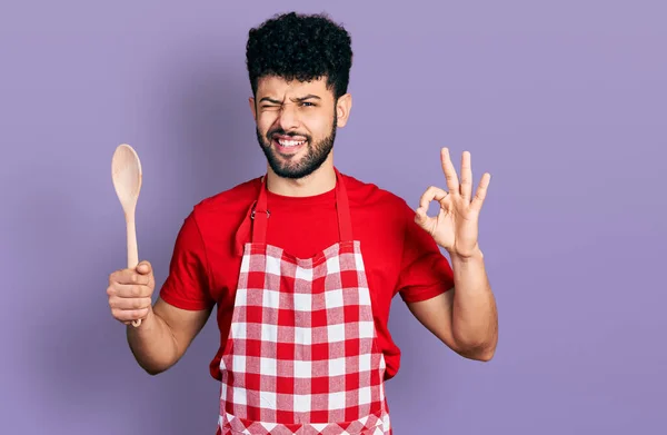 Young arab man with beard wearing baker uniform holding wooden spoon winking looking at the camera with sexy expression, cheerful and happy face. — Stock Image Young Arab Man Beard Wearing Baker Uniform Holding Wooden Spoon Stock Photo