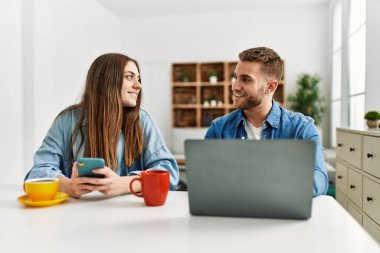 Genç beyaz çift kahvaltılarını yapıyorlar ve evde dizüstü bilgisayar ve akıllı telefon kullanıyorlar..