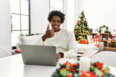 Genç Afro-Amerikalı adam, Noel ağacının yanında dizüstü bilgisayar kullanıyor ve elleriyle mutlu bir jest yapıyor. Kameraya bakıp başarıyı gösteren ifade. 