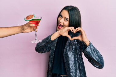 Seksi parti ceketi giymiş genç bir kız ve İspanyol kollu bir kokteyl sunuyor. Sevgiyle gülümseyen, elleriyle kalp sembolü yapan bir kız. romantik kavram. 