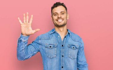 Günlük kot ceket giyen genç İspanyol adam beş numarayı gösterip işaret ederken kendinden emin ve mutlu gülümsüyor.. 