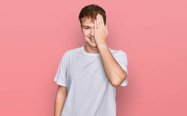 Beyaz günlük tişört giyen genç beyaz bir adam bir gözünü eliyle kapatıyor, yüzünde kendine güvenen bir gülümseme ve sürpriz bir duygu var.. 