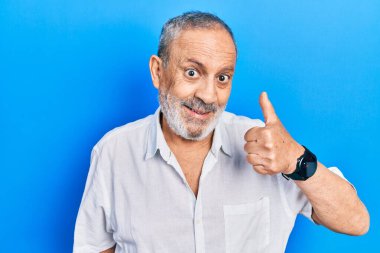 Sakallı, sıradan beyaz gömlekli yakışıklı bir adam mutlu bir el hareketi yapıyor. Kameraya bakıp başarıyı gösteren ifade. 