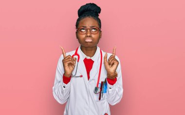 Doktor üniforması ve steteskop giyen genç Afro-Amerikan kadın üzgün ve üzgün görünüyor, parmaklarıyla yönü gösteriyor, mutsuz ve depresif.. 