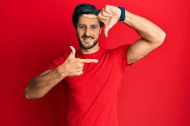Gündelik kırmızı tişört giyen genç İspanyol adam gülümseyerek elleri ve parmakları gülen yüzle çerçeve yapıyor. yaratıcılık ve fotoğrafçılık kavramı. 