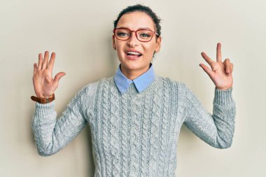 Kafkasyalı genç bir kadın günlük kıyafetler ve gözlük takıyor. Sekiz numaralı parmaklarıyla işaret ediyor. Kendinden emin ve mutlu bir şekilde gülümsüyor.. 