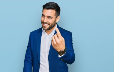 İş ceketi giyen genç İspanyol adam seni buraya çağırıyor. El kol hareketi yap. Mutlu bir şekilde karşıla ve gülümse. 