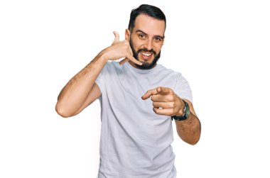 Young man with beard wearing casual white t shirt smiling doing talking on the telephone gesture and pointing to you. call me. 