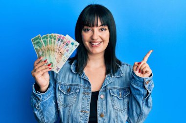 Young hispanic woman holding united arab emirates dirham banknotes smiling happy pointing with hand and finger to the side 