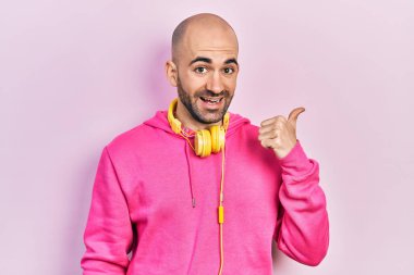 Young bald man wearing gym clothes and using headphones smiling with happy face looking and pointing to the side with thumb up. 
