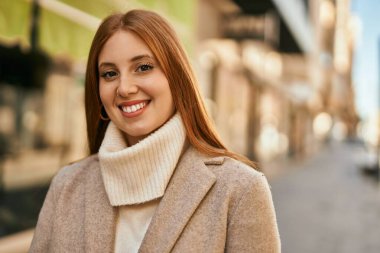 Genç kızıl kız gülümsüyor, mutlu bir şekilde şehirde duruyor..