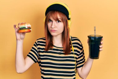 Burger yiyen, gazoz içen, kulaklık takan, kuşkucu ve gergin genç bir kadın sorun yüzünden surat asıyor. negatif kişi. 