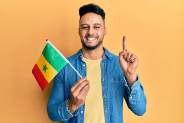 Young arab man holding senegal flag smiling with an idea or question pointing finger with happy face, number one 