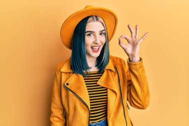 Young modern girl wearing yellow hat and leather jacket smiling positive doing ok sign with hand and fingers. successful expression. 