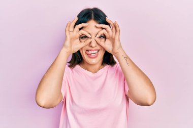 Young hispanic woman wearing casual pink t shirt doing ok gesture like binoculars sticking tongue out, eyes looking through fingers. crazy expression. 