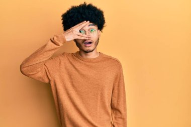 Young african american man with afro hair wearing casual winter sweater peeking in shock covering face and eyes with hand, looking through fingers with embarrassed expression. 