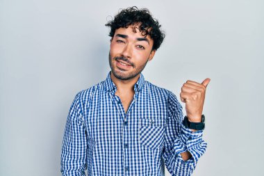 Gündelik kıyafetler giyen, gülümseyen, gülümseyen ve başparmağıyla yan tarafı gösteren İspanyol genç adam.. 