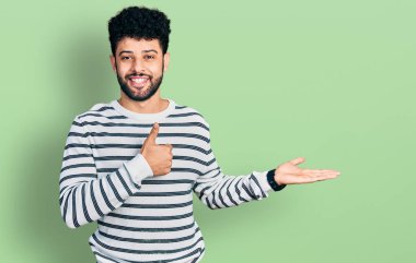 Sakallı, sıradan çizgili süveter giyen, avuç içi gösteren ve baş parmağıyla hareket eden mutlu ve neşeli gülümseyen genç bir Arap. 