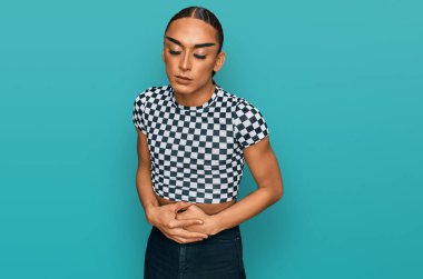 Hispanic transgender man wearing make up and long hair wearing modern clothes with hand on stomach because indigestion, painful illness feeling unwell. ache concept. 