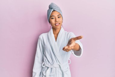 Young chinese woman wearing shower towel cap and bathrobe smiling friendly offering handshake as greeting and welcoming. successful business. 
