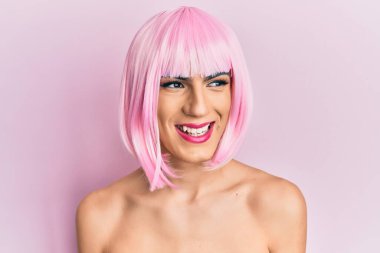 Young man wearing woman make up wearing pink wig looking away to side with smile on face, natural expression. laughing confident. 