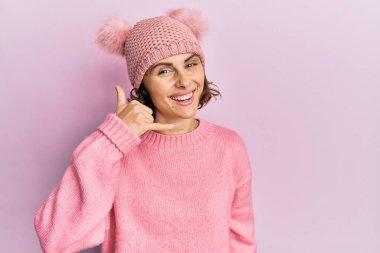 Genç esmer, sevimli yün şapkalı, gülümseyen, el ve parmaklarıyla telefon hareketi yapan, telefonda konuşan bir kadın. iletişim kavramları. 