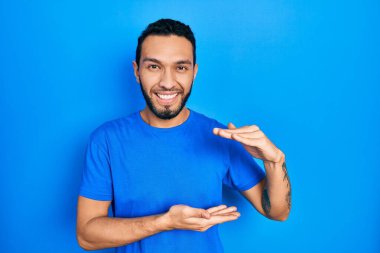 Hispanic man with beard wearing casual blue t shirt gesturing with hands showing big and large size sign, measure symbol. smiling looking at the camera. measuring concept. 