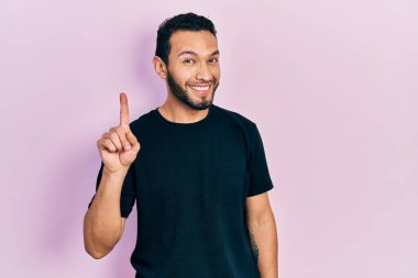 Hispanic man with beard wearing casual black t shirt smiling with an idea or question pointing finger up with happy face, number one 