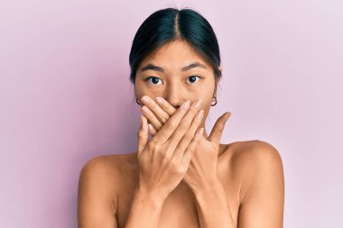 Young chinese woman standing topless showing skin shocked covering mouth with hands for mistake. secret concept. 