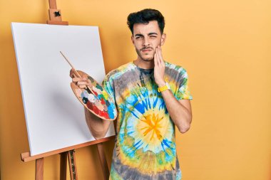 İspanyol asıllı genç bir adam, diş ağrısı ya da diş ağrısı yüzünden palet üstüne palet çizerek ayakta dikiliyor ve acı dolu bir ifadeyle ağıza dokunuyor. dişçi kavramı. 