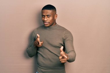 Balıkçı yaka kazak giyen genç siyahi adam mutlu ve komik yüzlü parmaklarını kameraya doğrultuyor. iyi enerji ve titreşimler. 