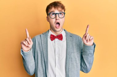 Young caucasian nerd man wearing glasses wearing hipster elegant look with bowtie amazed and surprised looking up and pointing with fingers and raised arms. 