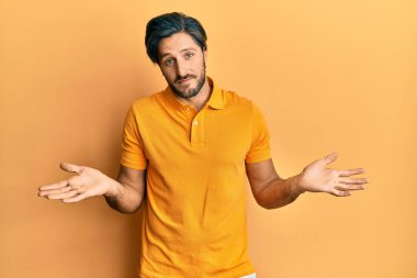 Sıradan sarı tişörtler giyen, kolları ve elleri havada, kafası karışık, genç İspanyol bir adam. şüphe kavramı. 