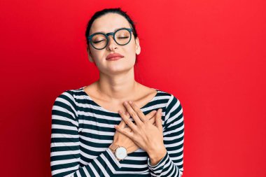 Gündelik kıyafetler ve gözlük takan genç beyaz bir kadın elleri göğsünde, gözleri kapalı ve yüzünde minnet dolu bir jest ile gülümsüyor. sağlık kavramı. 