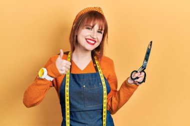 Kızıl saçlı genç kadın terzinin atölye önlüğü giymiş elinde makas, mutlu ve pozitif gülümsüyor, başparmağıyla mükemmel ve onay işareti yapıyor. 