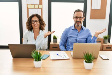 Orta yaşlı İspanyol kadın ve erkek ofiste dizüstü bilgisayarıyla oturmuş neşeli bir şekilde sunum yapıyor ve avuç içi kameraya bakıyor.. 