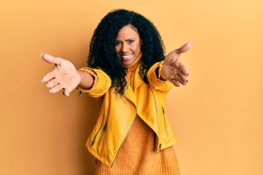 Orta yaşlı Afro-Amerikalı kadın yün kazak ve deri ceket giyip kameraya bakıyor ve kucaklaşmak için gülümsüyor. Mutluluğu kucaklayan neşeli bir ifade.. 