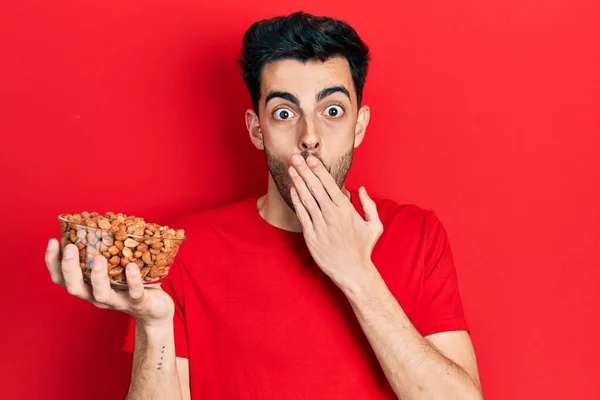 Joven Hispano Sosteniendo Cacahuetes Cubriendo Boca Con Mano Sorprendido Temeroso — Foto de Stock